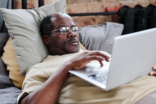 Restful African American male pensioner in eyeglases and t-shirt using laptop for communication while lying in bed after sleep in the morning