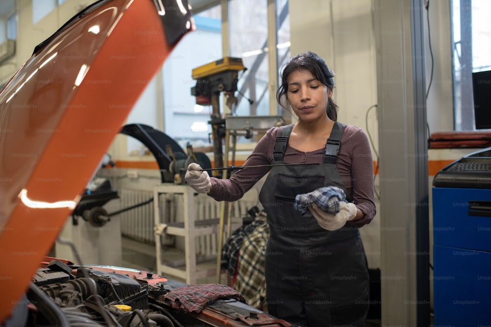 Porträt einer jungen Mechanikerin, die den Ölstand mit einem Ölmessstab überprüft, während sie das Auto in der Werkstatt repariert, Kopierraum