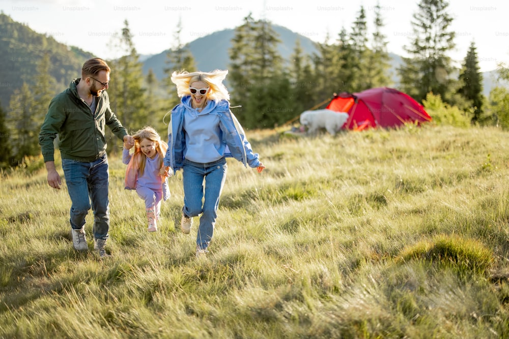 Young caucasian couple with little girl and dog have fun, running together while travel with tent in the mountains. Happy family summer vacation at campsite