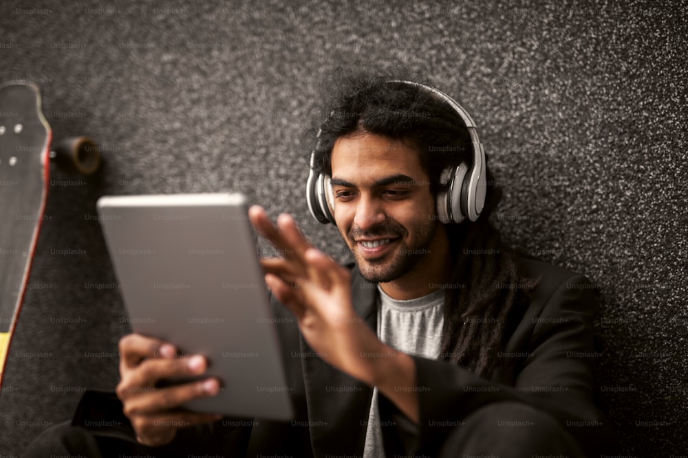 Elegante giovane hipster dreadlock con le cuffie seduto appoggiato al muro grigio e pattina vicino a lui usando un tablet con un sorriso.