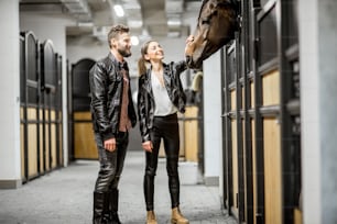美しい厩舎に立って馬の頭を撫でる革ジャンを着た若いカップルの騎手