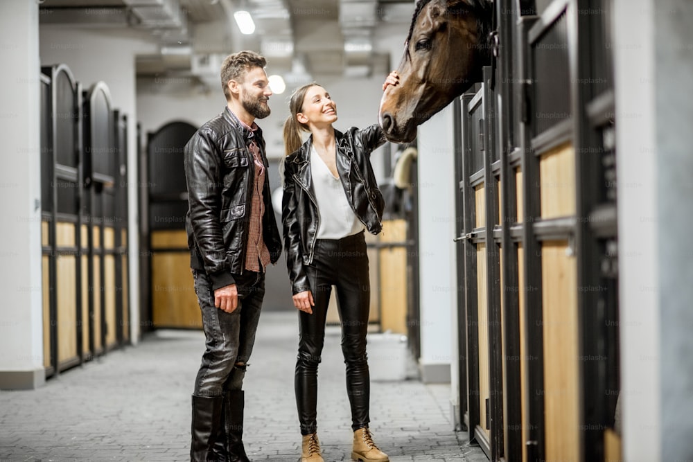 Young couple riders in leather jackets standing in the beautiful stable stroking a horse head