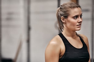 Jeune femme blonde en forme dans un débardeur sportif à l’air concentré tout en se tenant seule à la salle de sport avant une séance d’entraînement