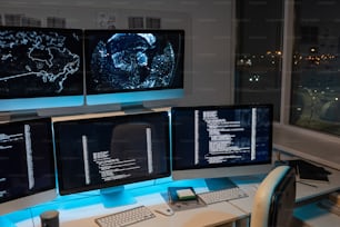 Part of large openspace office of programming company with long desk and several computer monitors with coded data