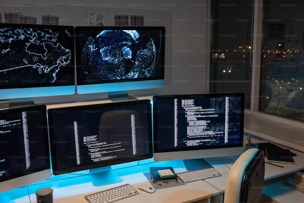 Part of large openspace office of programming company with long desk and several computer monitors with coded data