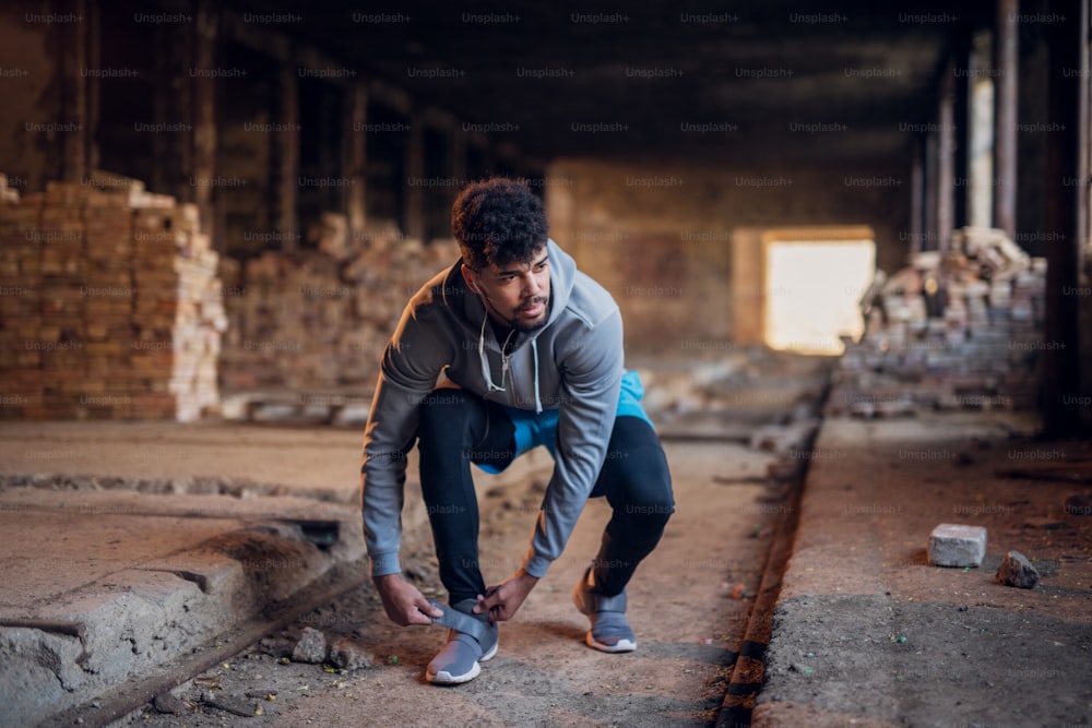 Close up view of sporty active afro american runner man crouching and tying velcro sneakers in the abandon place.