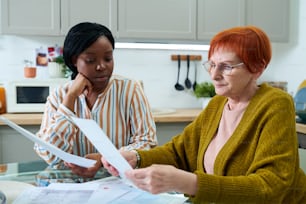 Senior woman in eyeglasses examining financial documents at table in kitchen together with social worker