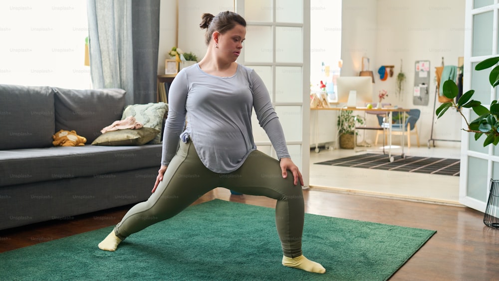 Young female with Down syndrome doing physical exercise for stretching legs while standing on green carpet in living room during workout