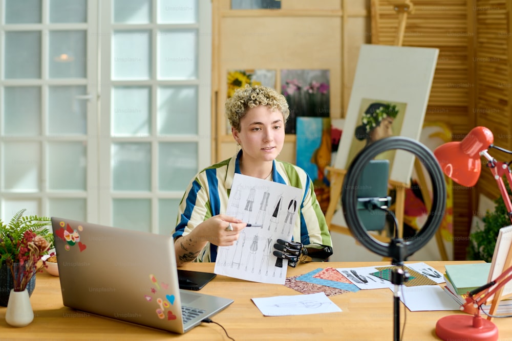 Young female fashion designer with arm prosthesis showing sketch of new seasonal collection of clothes to online audience