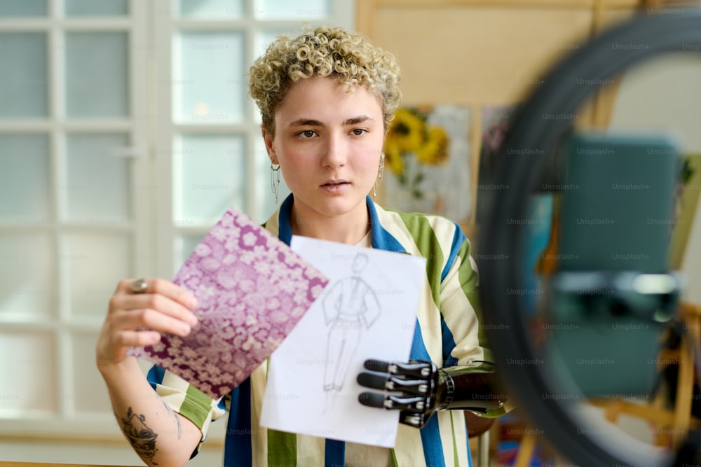 Young creative female fashion designer showing drawing of new model for seasonal collection to online audience during training