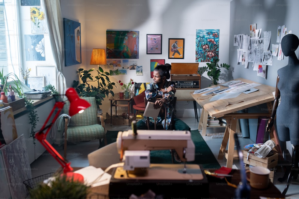 Plano largo de un joven artista moderno y diseñador de ropa de etnia india sentado en una silla en la sala de estar de su casa mirando hacia otro lado