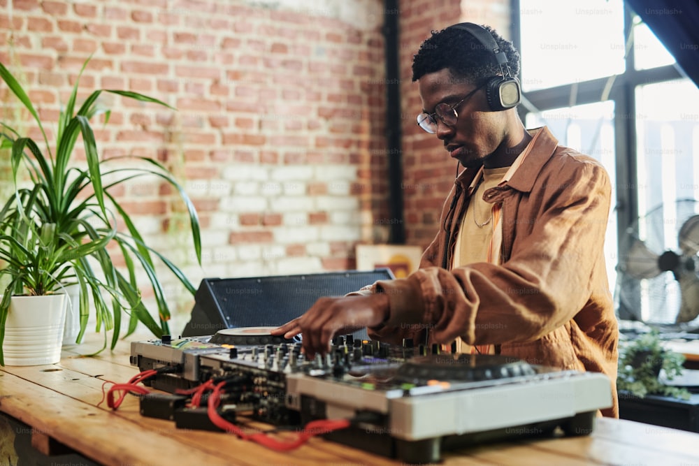 Ernsthafter junger schwarzer Mann mit Kopfhörern auf dem Kopf, der Geräusche mischt, während er an Plattenspielern steht und im Studio Musik macht
