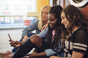 Diversas amigas jóvenes riendo mientras están sentadas juntas en el piso de una lavandería usando un teléfono celular