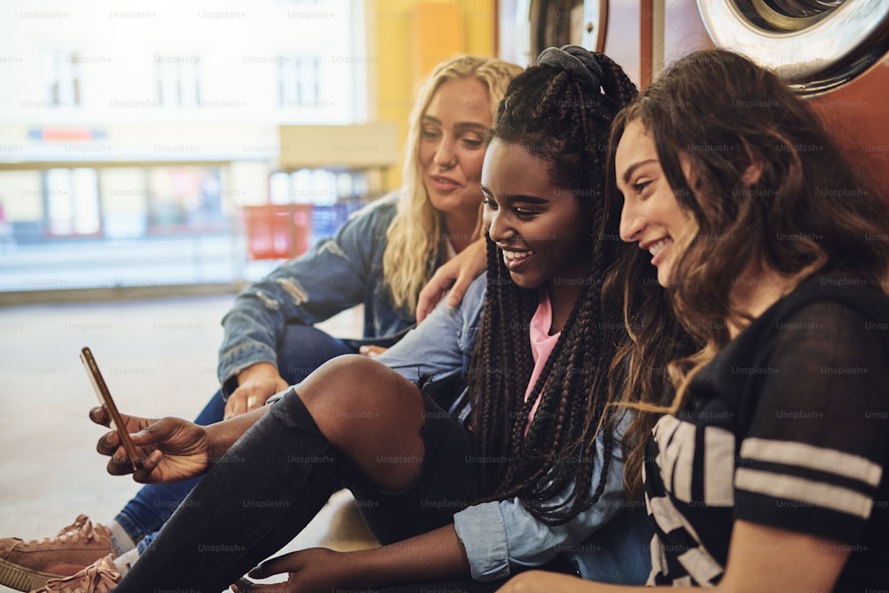 Verschiedene junge Freundinnen lachen, während sie zusammen auf dem Boden eines Waschsalons sitzen und ein Handy benutzen