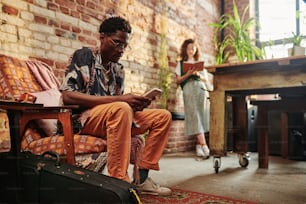 Young serious black man in casualwear using mobile phone while sitting in armchair against his girlfriend reading book while standing by wall
