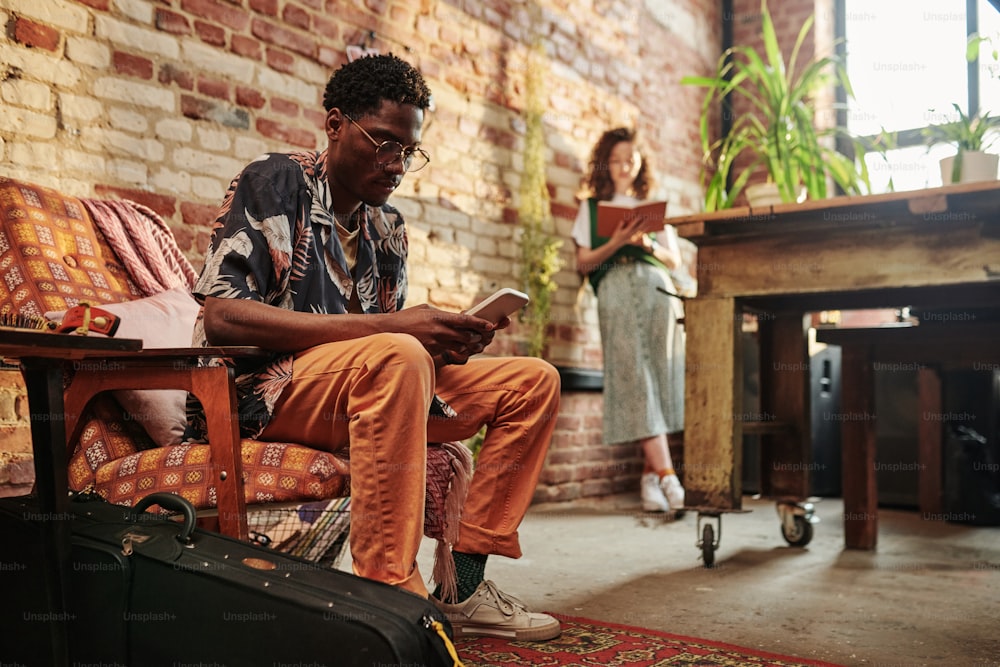 カジュアルウェアの若い真面目な黒人男性が、壁際に立って本を読んでいるガールフレンドに対して肘掛け椅子に座りながら携帯電話を使用しています