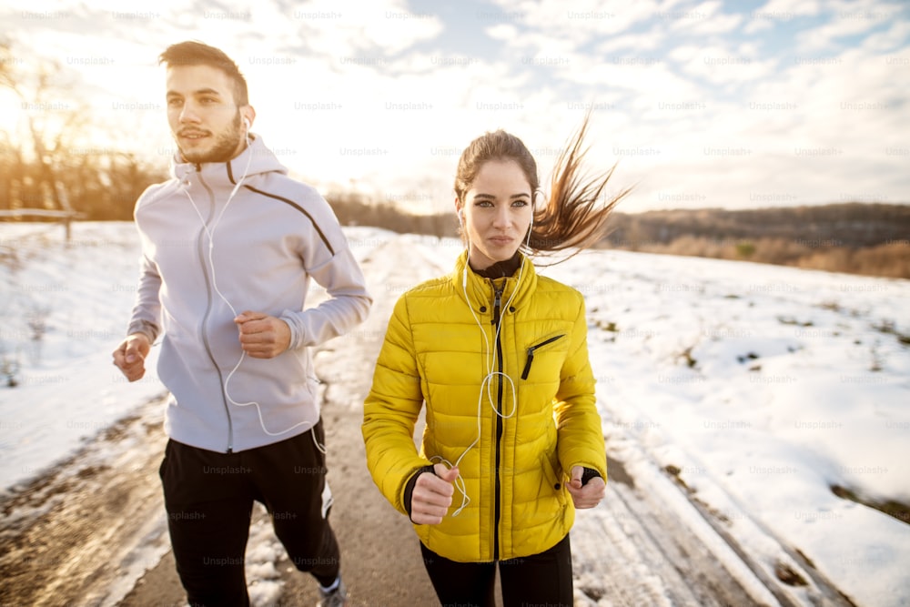 Portrait view of young beautiful satisfied motivated and focused fitness love couple in winter sportswear running in the snowy nature with earphones.
