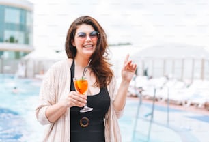 Stylish happy young woman forty years plus size body positive in black swimsuit and sunglasses enjoying her life drinking with glass of cocktail near hotel pool