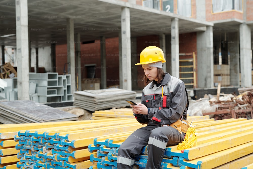 Mujer joven en ropa de trabajo sentada en una pila de tablones amarillos y enviando mensajes de texto en el teléfono inteligente mientras descansa después del trabajo en el sitio de construcción