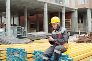Giovane donna in abbigliamento da lavoro che si siede su una pila di assi gialle e invia messaggi di testo nello smartphone mentre ha una pausa dopo il lavoro in cantiere