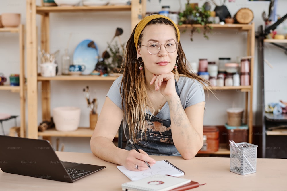 캐주얼웨어와 안경을 쓴 젊은 현대 여성이 스튜디오의 직장에 앉아 메모장에 스케치를 하면서 카메라를 보고 있다