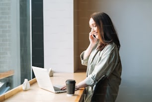 Femme d’affaires brune souriante adulte de quarante ans aux cheveux longs en chemise élégante travaillant sur un ordinateur portable à l’aide d’un téléphone portable dans un lieu public dans un café