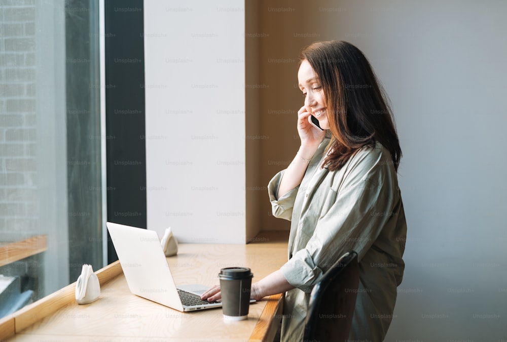 Adult smiling brunette business woman forty years with long hair in stylish shirt working on laptop using mobile phone at public place in cafe