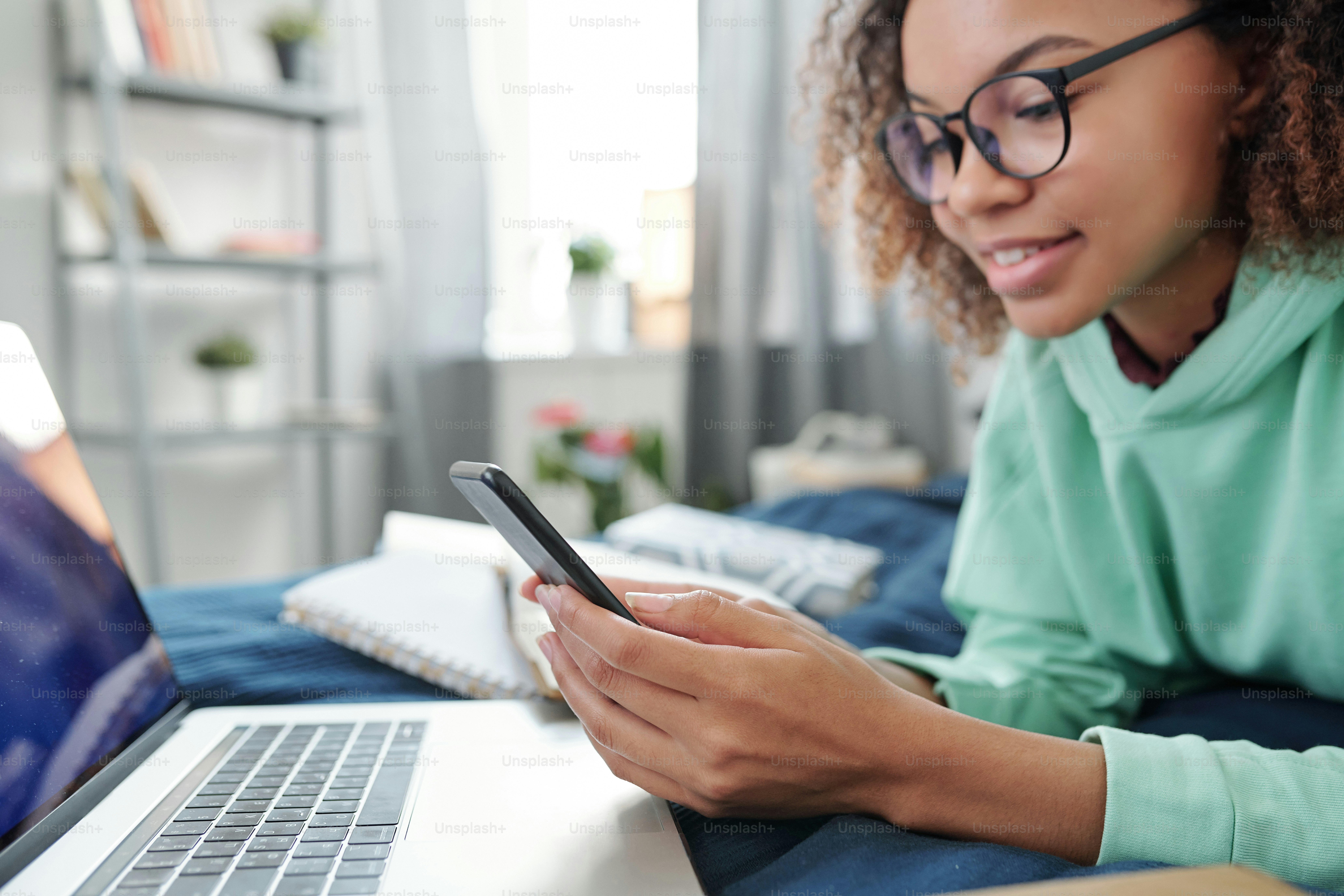 Wanita muda ras campuran berkacamata bergulir di smartphone sambil berbaring di tempat tidur di depan laptop dan mempersiapkan tugas di rumah