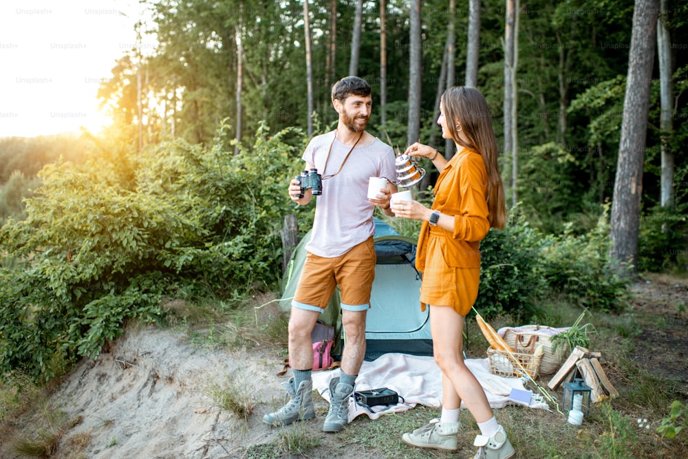 Young couple standing with hot drinks at the camping in the forest, enjoying their summer trip during the sunset