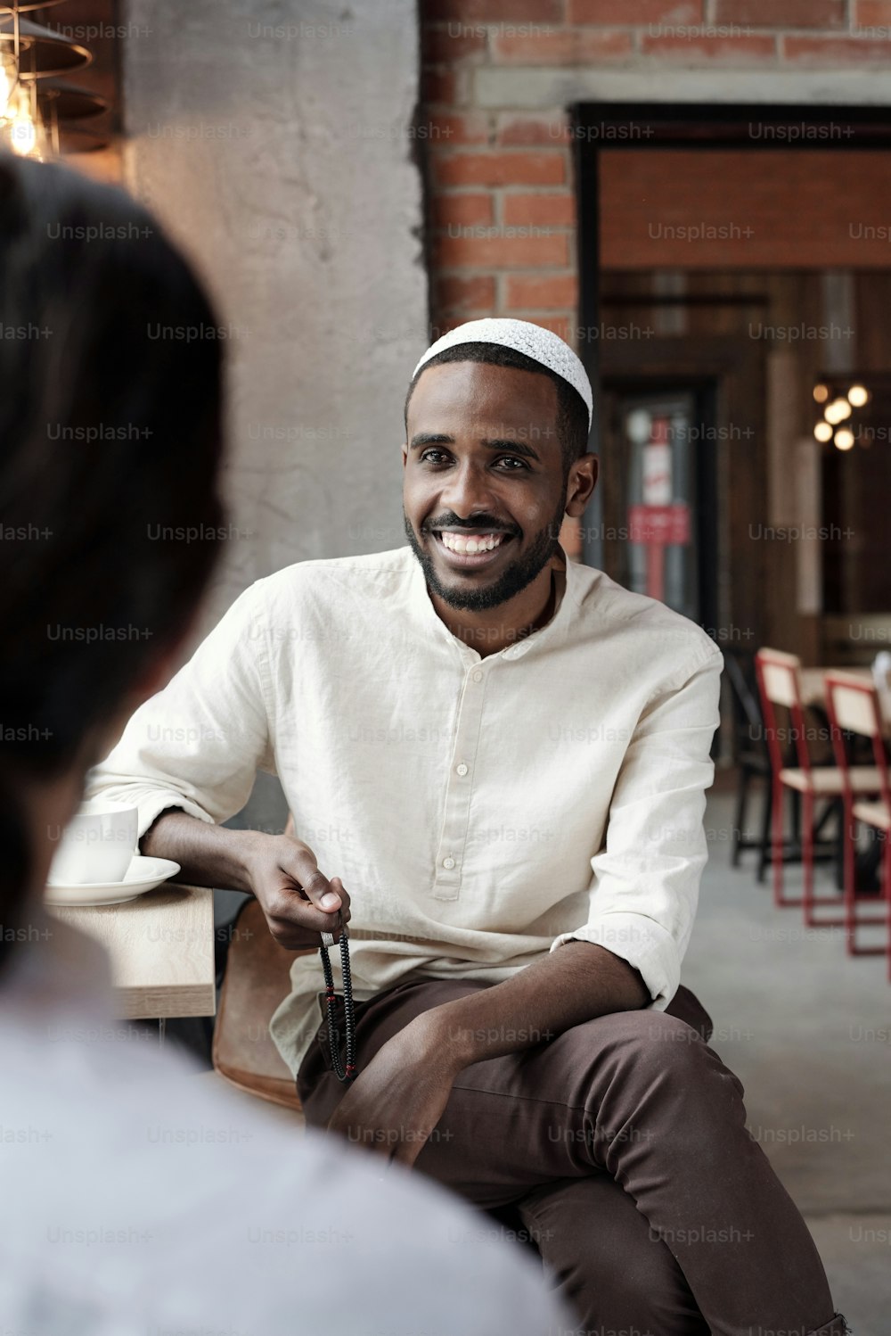 Positive young Black man in kufi cap sitting at table in loft cafe and talking to friend