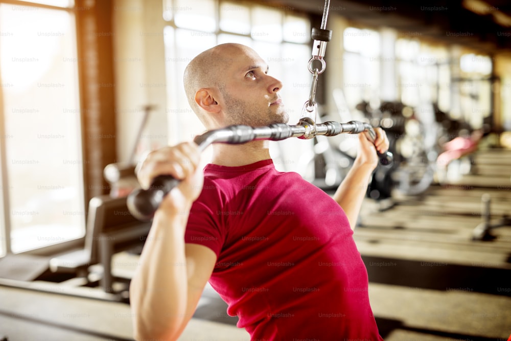 Close up side view of motivated and focused strong muscular active healthy young bald man sitting on the bench and pulling the metal bar from the machine for back muscles.