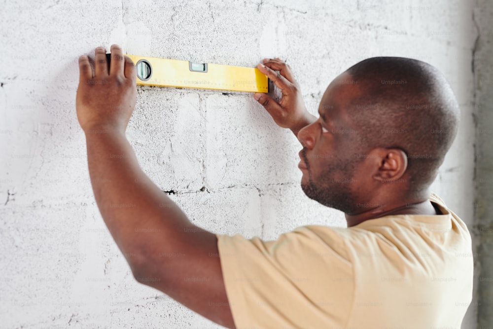 Young man of African ethnicity standing in front of white painted brick wall and using level measuring handtool during home renovation