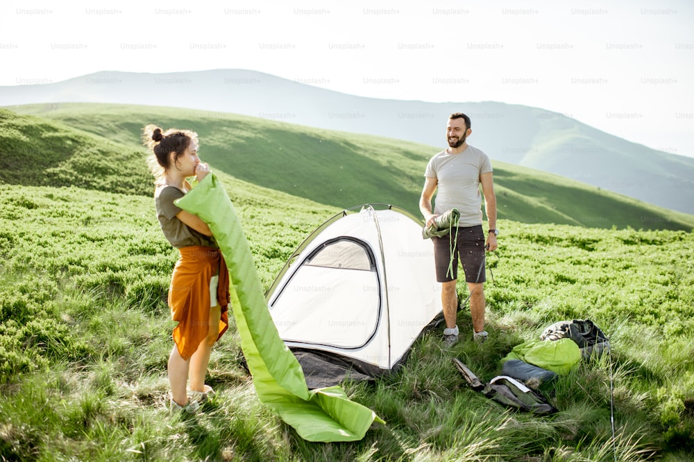 Young couple setting up the tent and inflating the mattress on the green meadow, traveling high in the mountains during the sunset