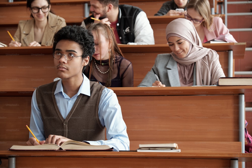 Contemporary intercultural university students making notes while sitting by long wooden desks in lecture hall and listening to professor at lesson