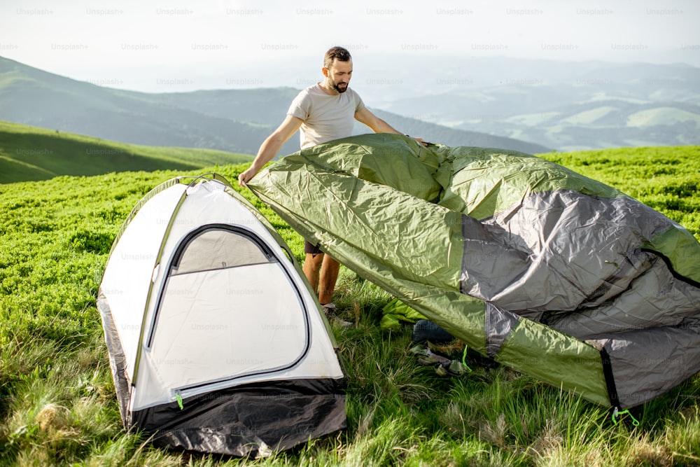 Man setting up the tent on the green meadow, traveling high in the mountains during the sunset