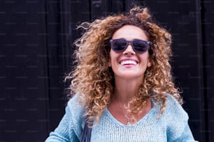 Joyful and happiness concept with cheerful beautiful adult caucasian woman smile and laugh in front of the camera and black wall in background - happy people portrait outdoor - long curly blonde hair