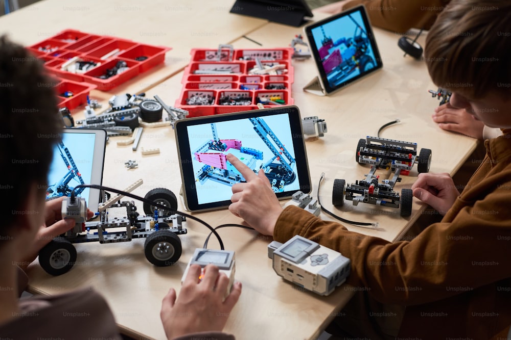 Vista posteriore del ragazzo che tocca lo schermo della tavoletta digitale che controlla il suo robot con il computer mentre si siede al tavolo alla lezione di robotica