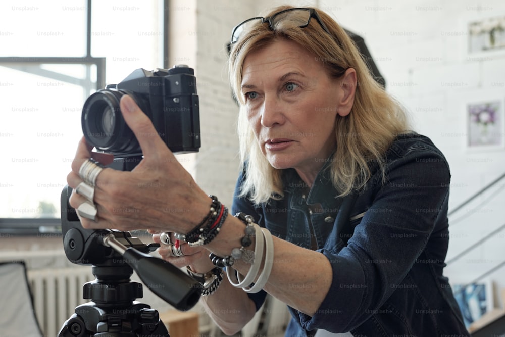 Dama madura concentrada con cabello rubio ajustando la lente de la cámara mientras toma fotos en un estudio moderno