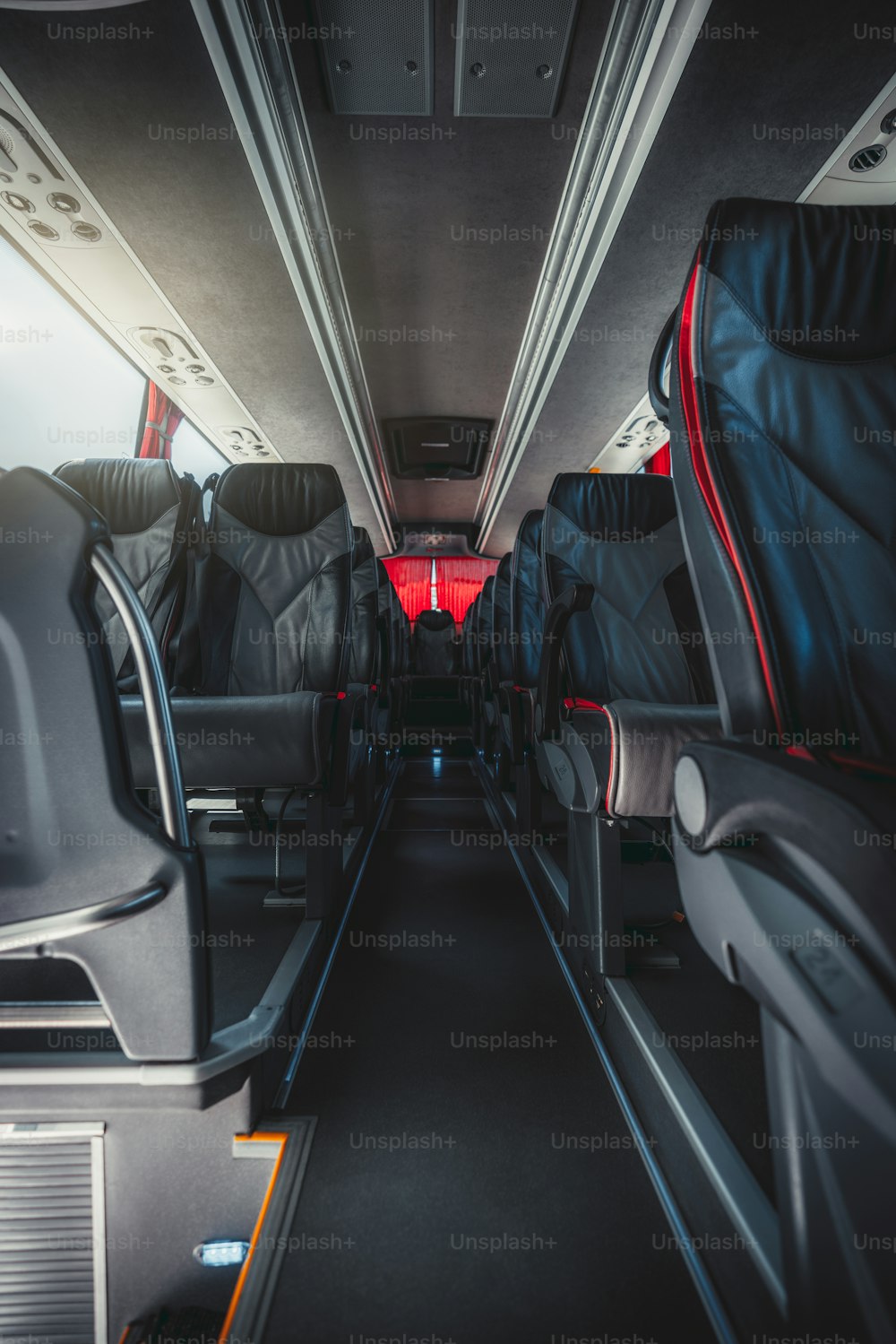 A vertical wide-angle view of an empty interior of a regular intercity bus with rows of leather numbered seats with red borders, carpeted seat aisle in the center