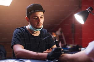 Young focused tattoo artist is inking customers arm carefully in his shop.