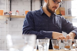 Portrait of man in glasses writing results of coffee cupping test, examining freshle ground coffee for flavour. He is standing near white wall in front of rows with glass cups