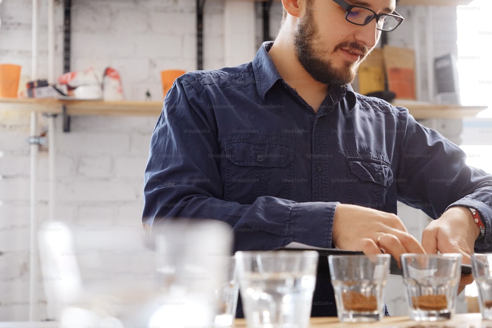 Retrato de un hombre con gafas escribiendo los resultados de la prueba de catación de café, examinando el café molido fresco para el sabor. Está parado cerca de una pared blanca frente a filas con vasos de vidrio