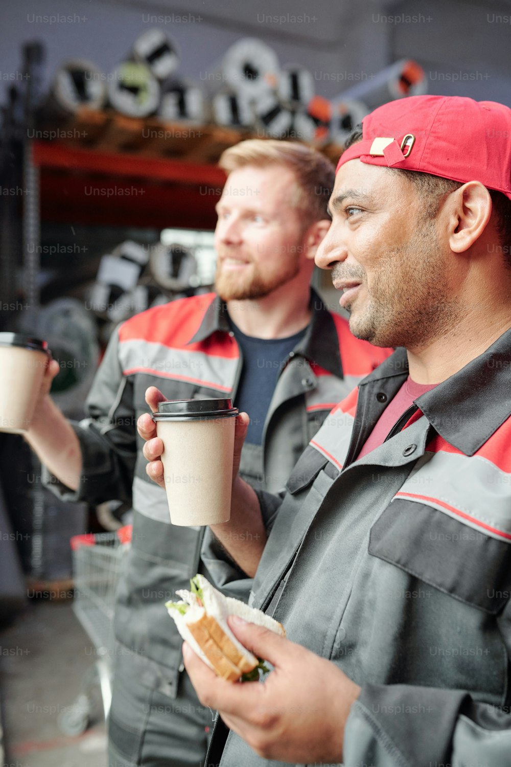 Zwei glückliche junge Lagerarbeiter mit Sandwiches und Kaffee schauen sich neue Arbeitsgeräte an, während sie in der Pause am Arbeitsplatz zu Mittag essen