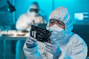 Joven mujer asiática en ropa de trabajo protectora mirando la placa de circuito en sus manos enguantadas mientras trabaja en el laboratorio