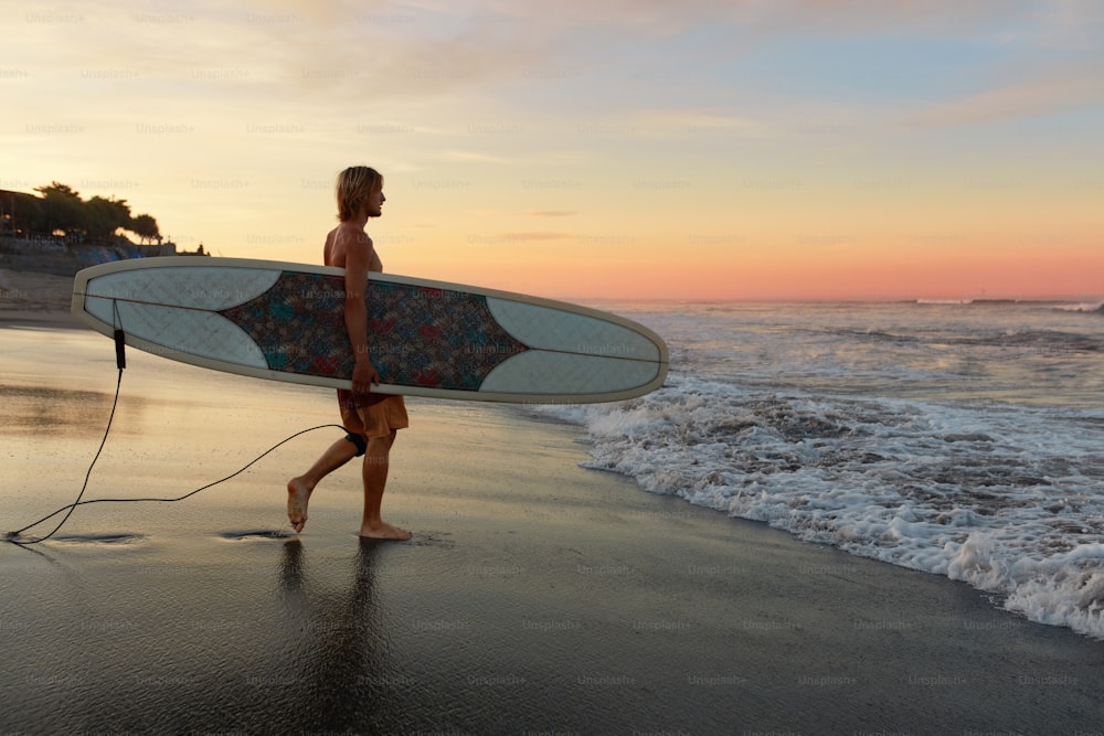 サーファー。砂浜を歩く白いサーフボードを持つサーフィンマン。アクティブなライフスタイルのためのウォータースポーツ。美しい海、海の夕日の景色。