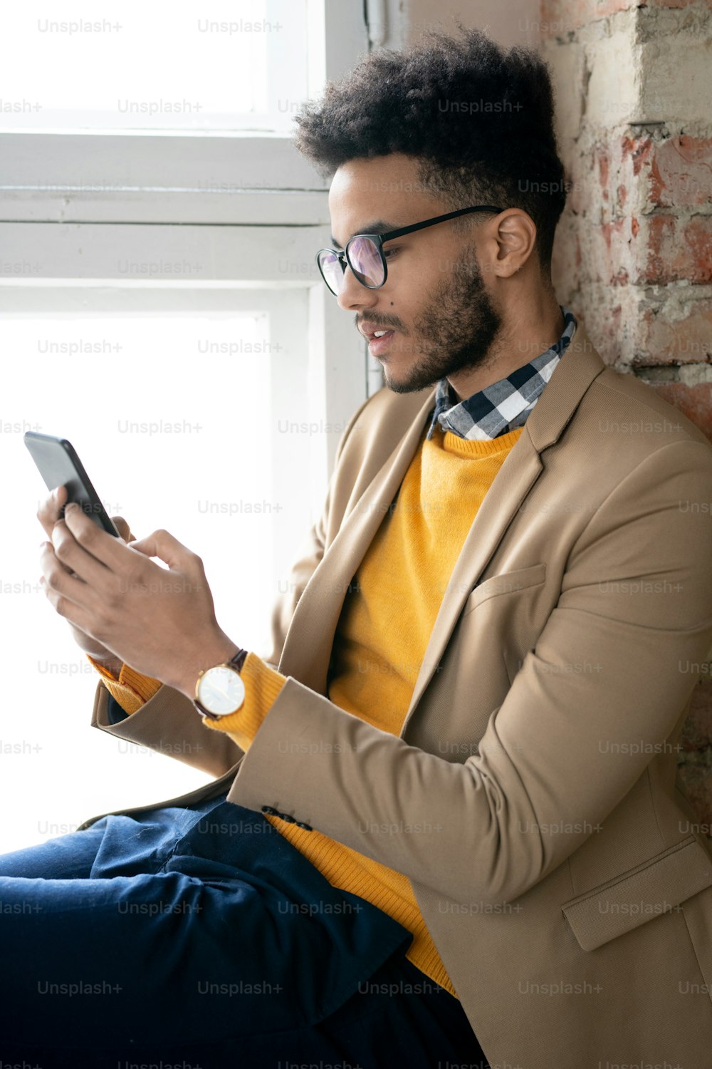Serious young bearded man in eyeglasses sitting on window sill and texting message on phone while working online