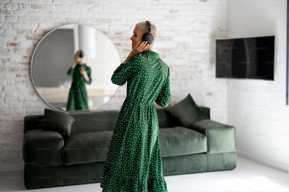 La mujer elegante con vestido verde disfruta de la música con auriculares y teléfono celular bailando en la sala de estar de casa. Concepto de vida confortable y tiempo libre