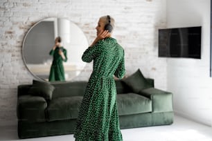 Stylish woman in green dress enjoys the music with headphones and cell phone dancing in the living room at home. Comfort living and leisure time concept