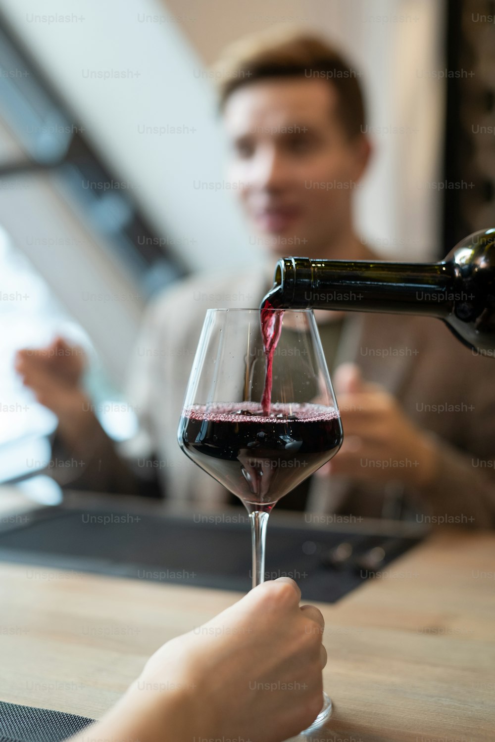 Hand of girl sitting by served table in front of her boyfriend during dinner and holding wineglass while waiter pouring red wine into it