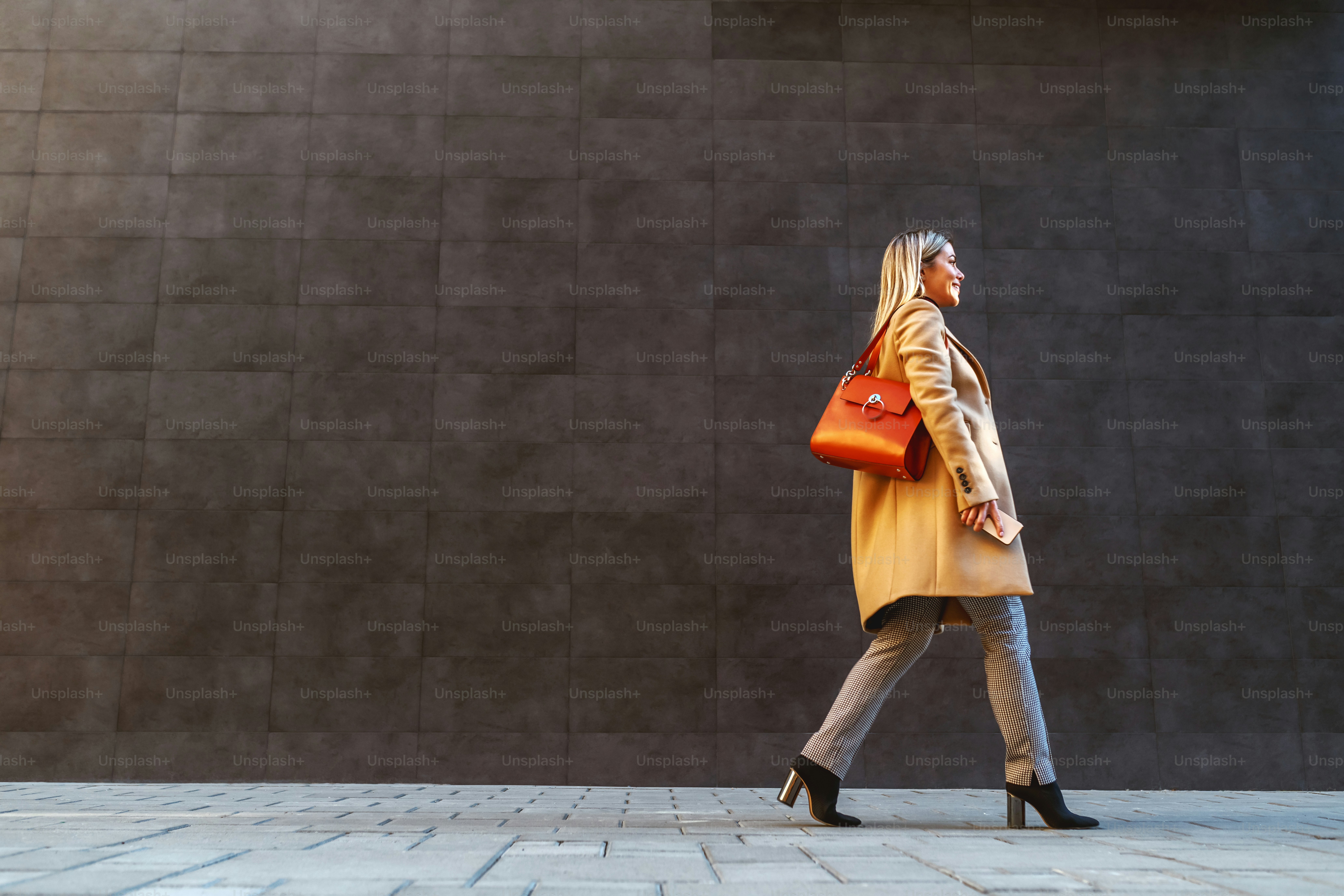 Side view of attractive caucasian blond woman in coat passing by gray wall outside.